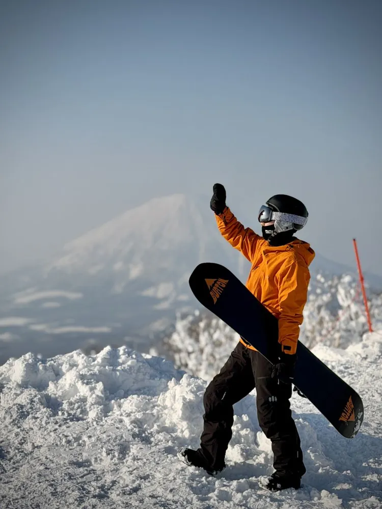 Hao Jun holding a snowboard on a snowy ridge, waving, with a mountain behind